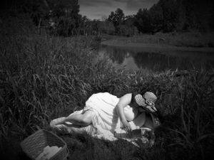 Moody landscape photograph with figure in white dress resting among tall grasses near water.