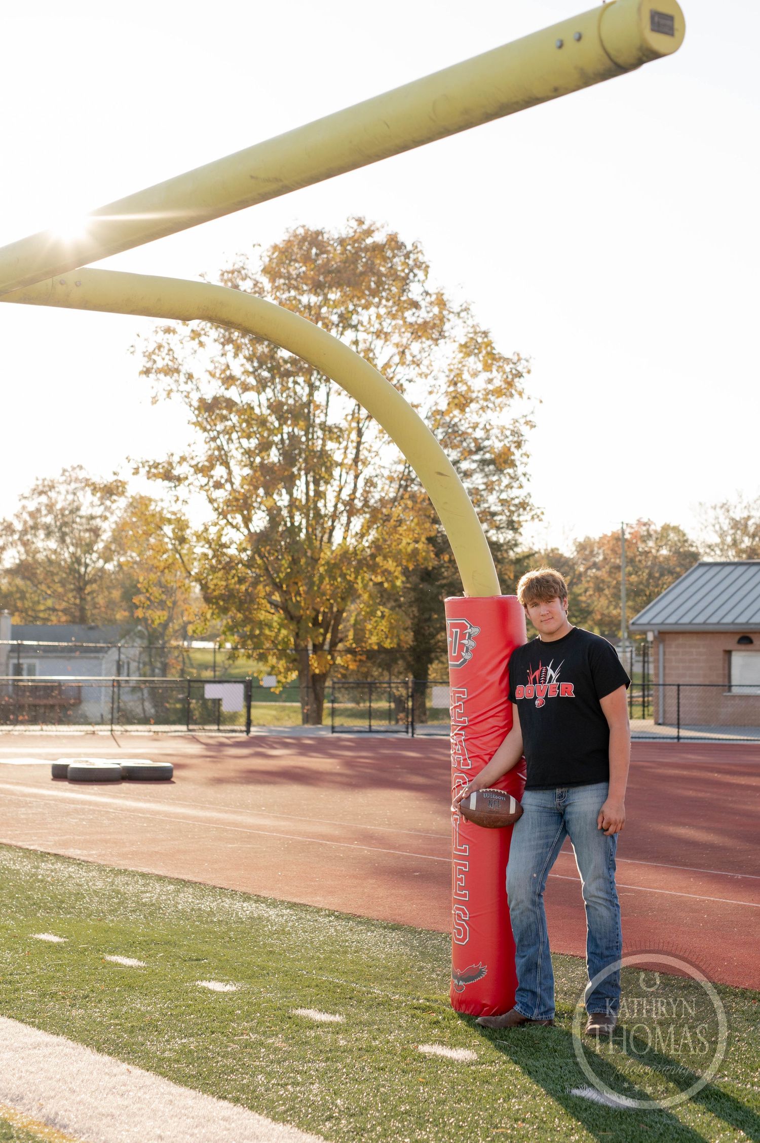 Player stands by goal post padding on football field with autumn trees in background.