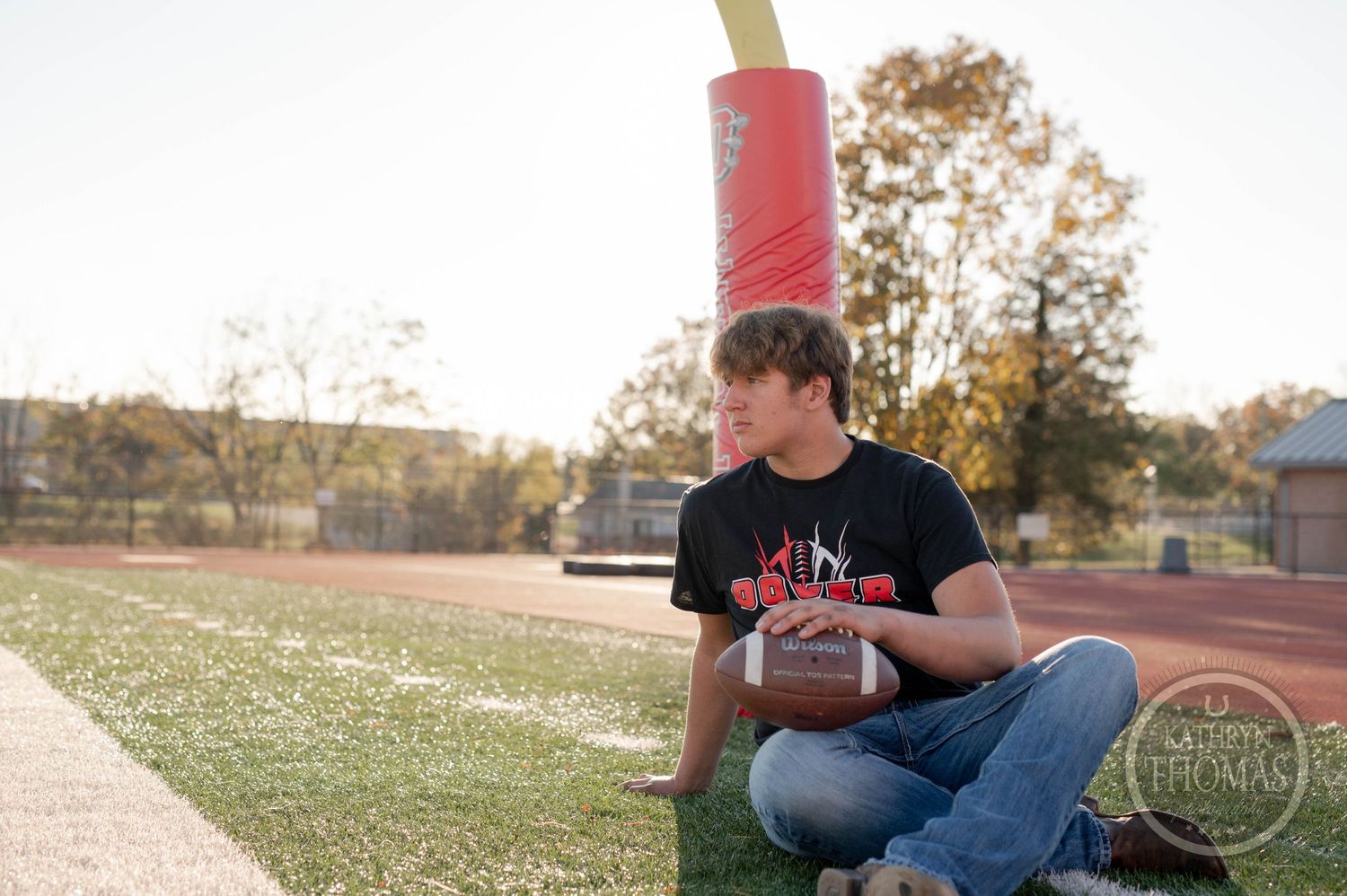 Player sits with football by goal post on field during golden hour lighting.