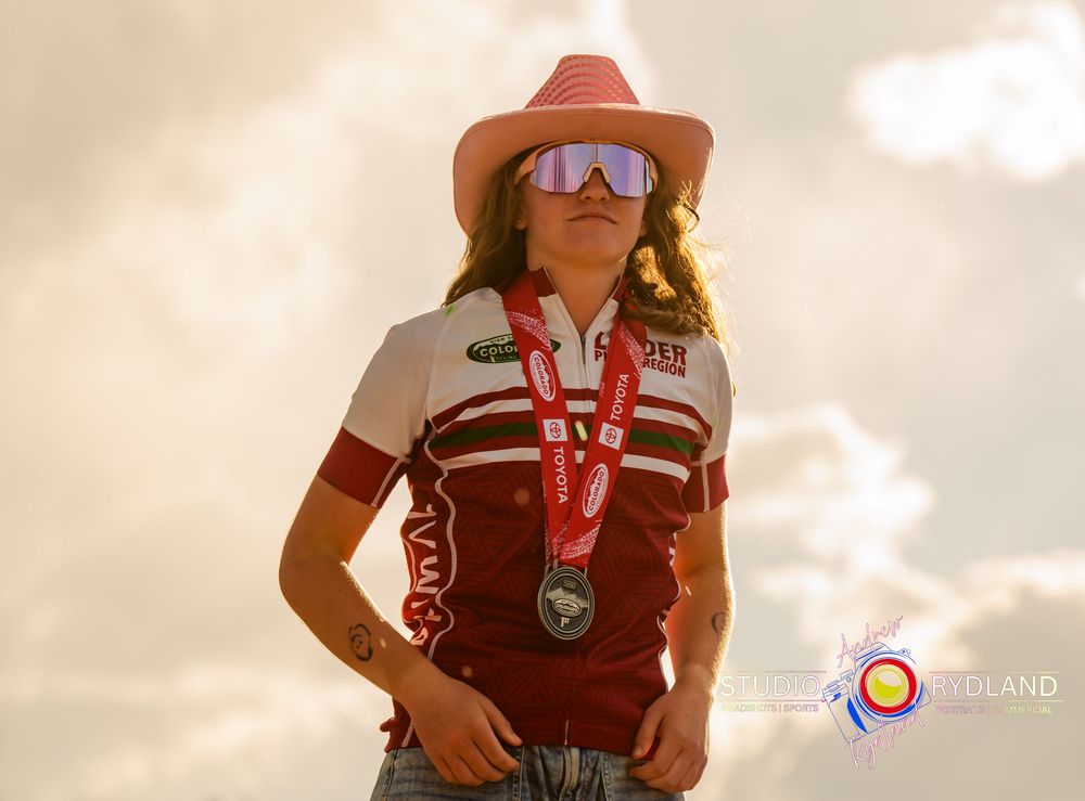 Athlete wearing brown hat, sunglasses and red jersey stands proudly with medal against cloudy sky at sunset.