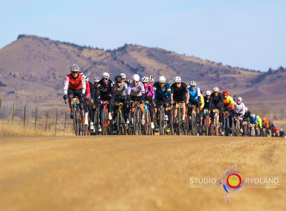 A panoramic view of cyclists riding in a group across a golden grassland with mountains in the background.