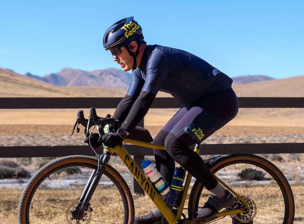 Cyclist in dark athletic gear riding a gravel bike through scenic desert mountain terrain on a sunny day.