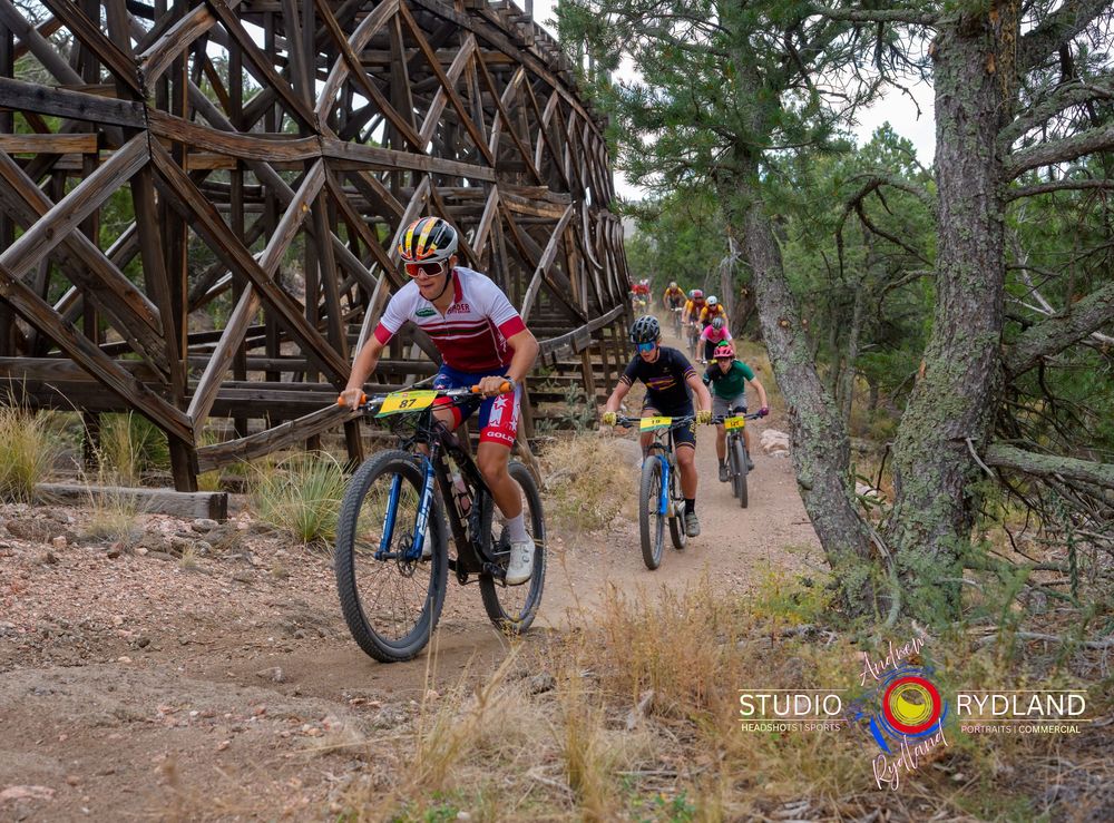 Mountain bikers riding on a dirt trail past a wooden bridge structure in a wooded area.