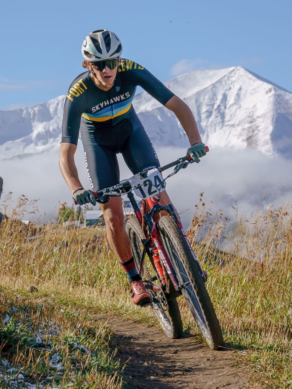 Cyclists riding mountain bikes on a dirt trail with snow-capped peaks in the background.
