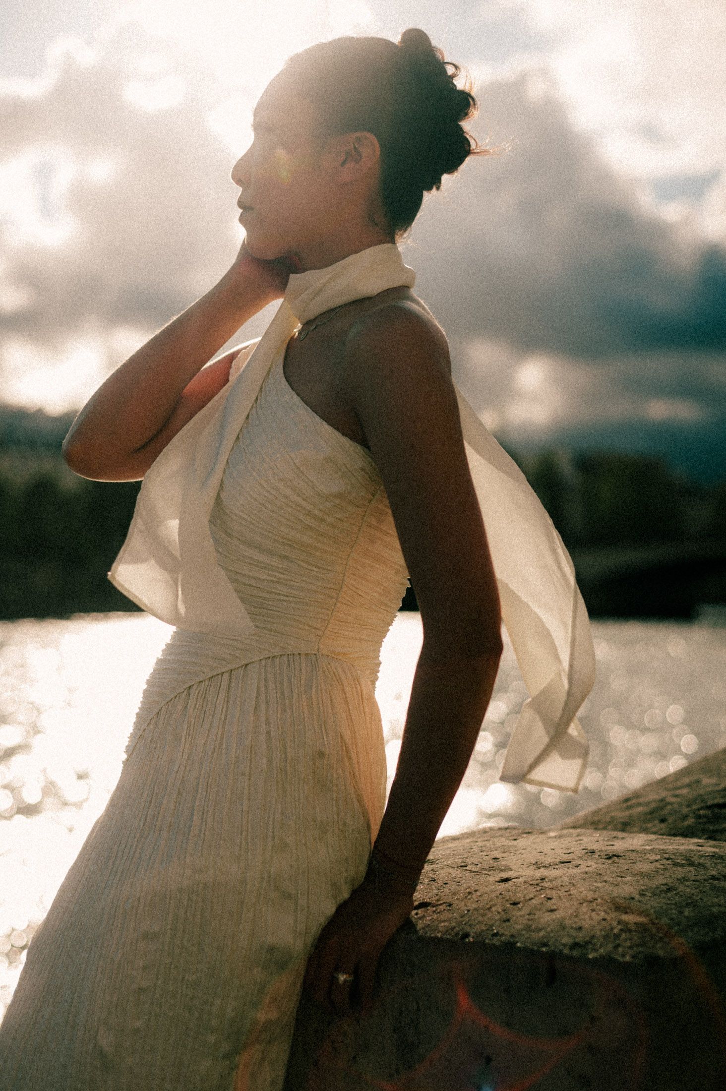 A person in a white dress poses against a dramatic cloudy sky during sunset.