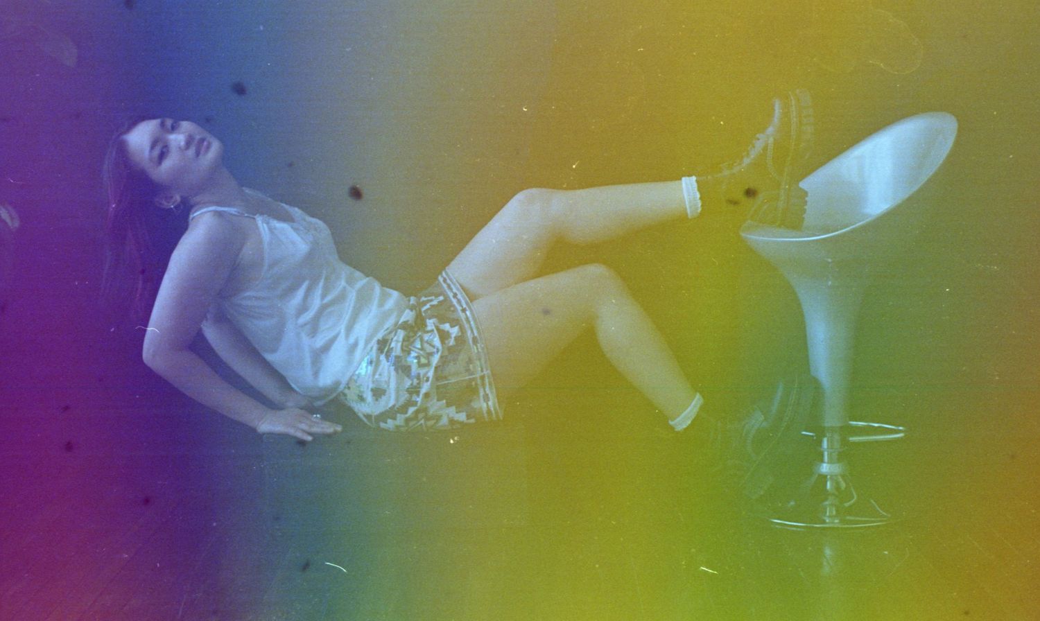 Artistic photograph with rainbow light leaks showing a reclining figure in vintage style.