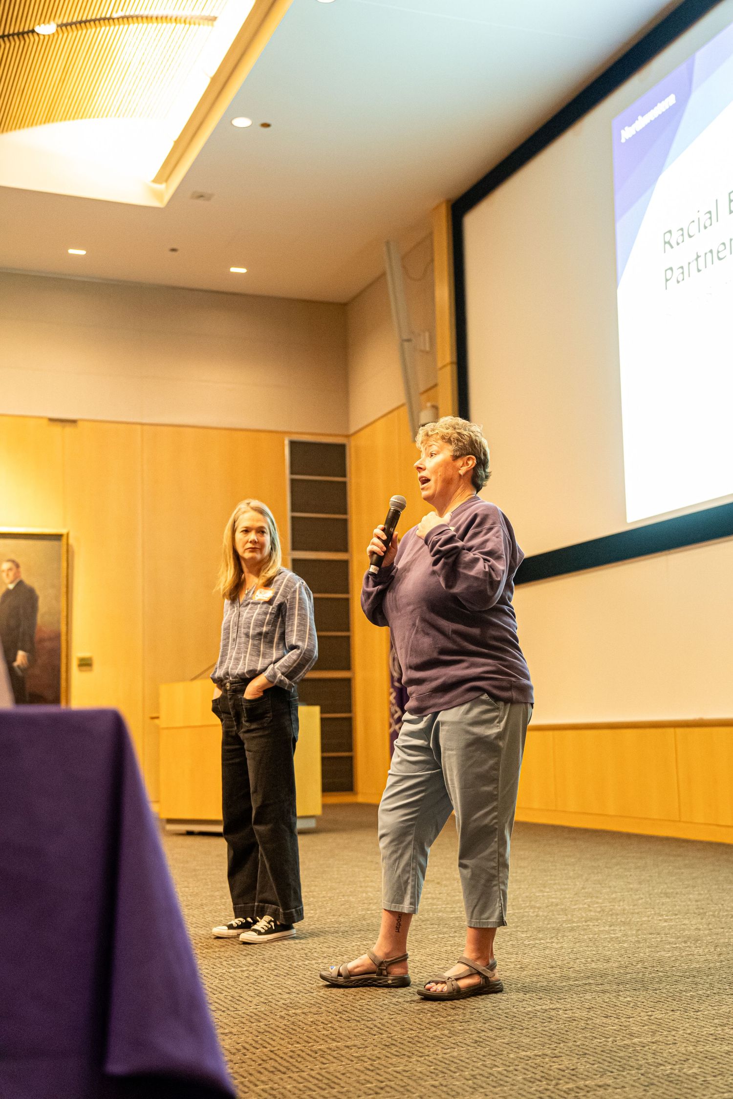 Conference presenters speaking at a podium in a modern lecture hall with wood paneling.