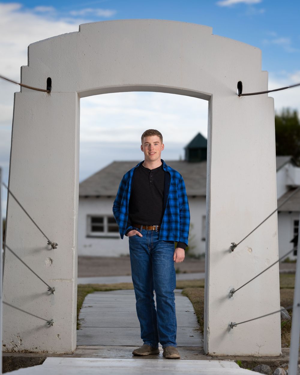 Casual portrait of someone wearing blue plaid flannel and jeans standing in a white archway entrance.