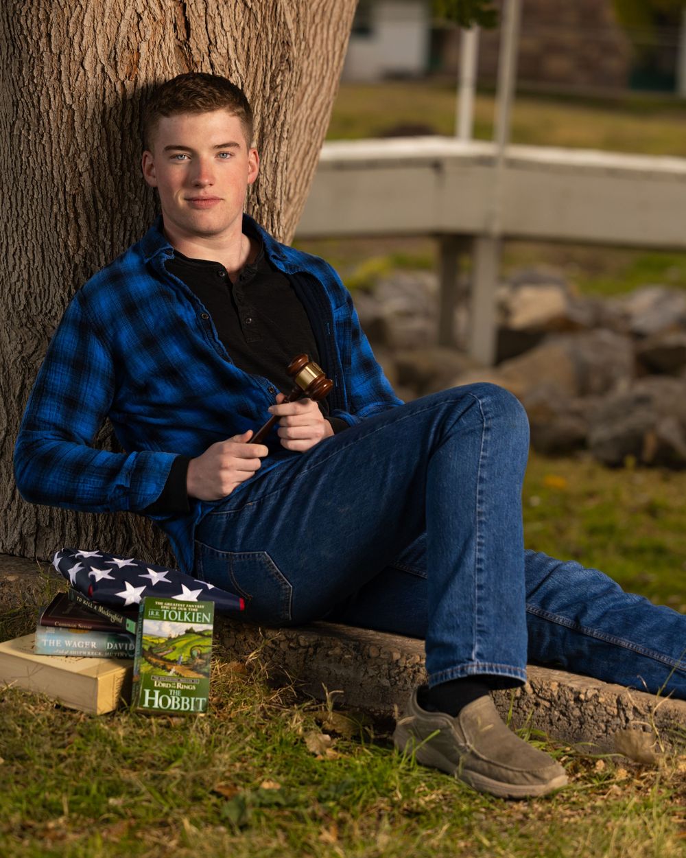 Casual outdoor portrait next to tree showing plaid shirt, jeans and boots with hobby items.