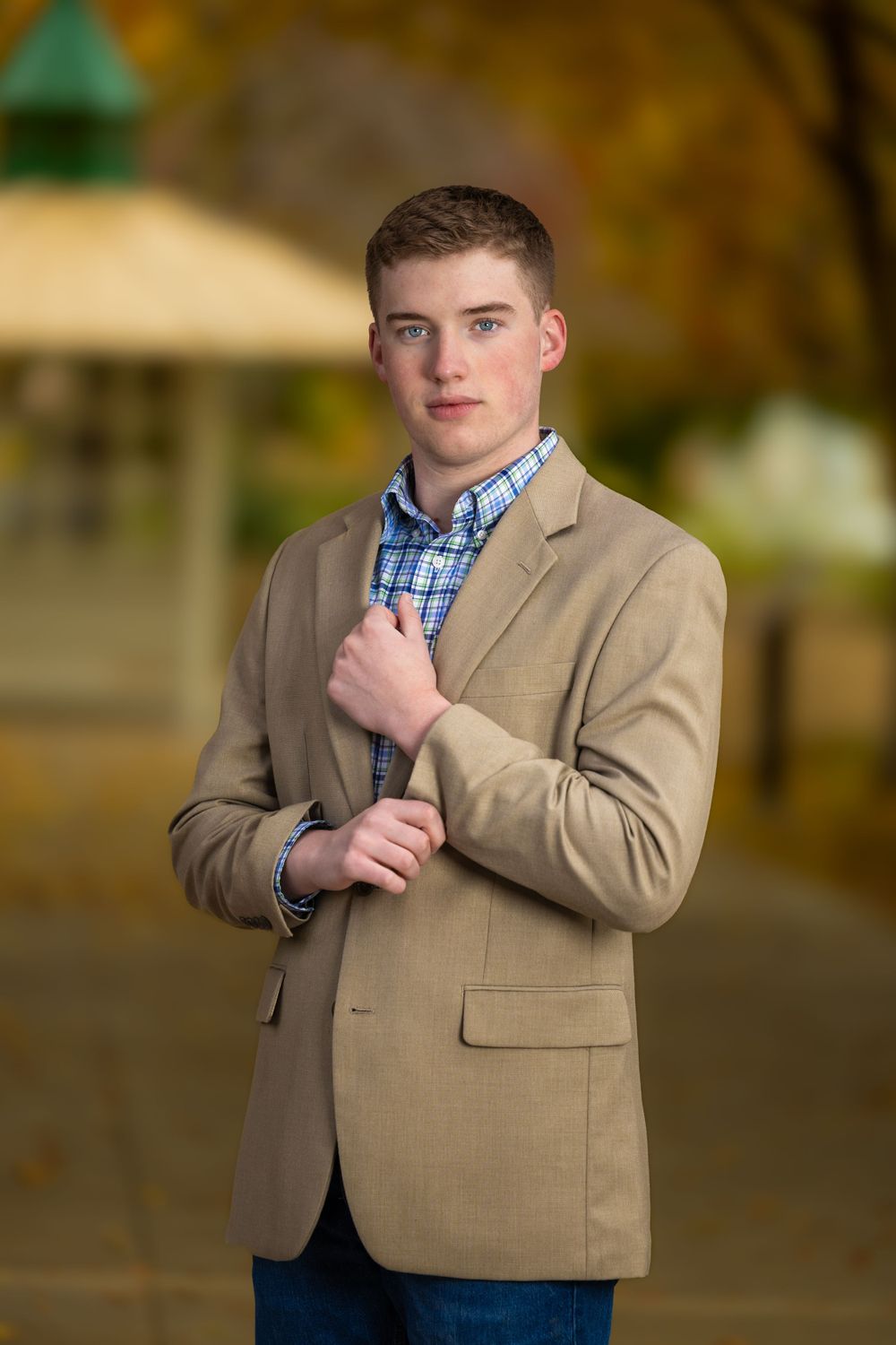 Professional portrait outdoors featuring khaki blazer and plaid button-up shirt against autumn background.