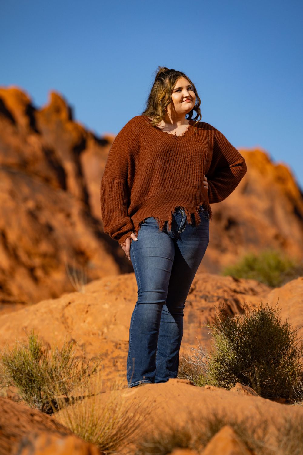 Person wearing brown sweater and jeans stands on red rock formations against bright blue sky.