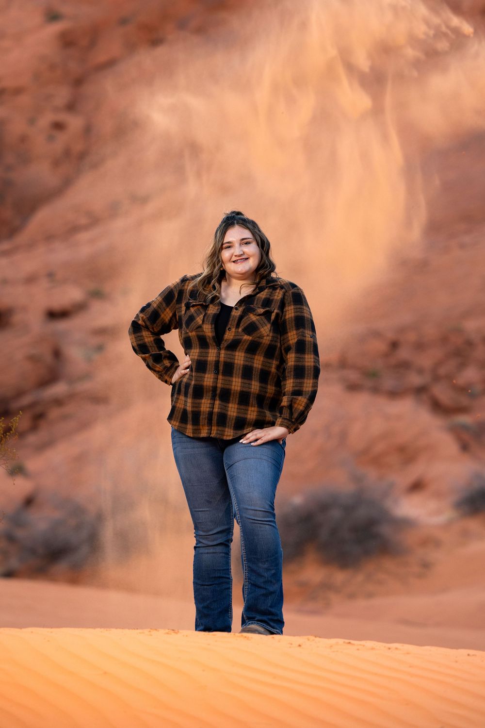 A woman wearing a plaid flannel jacket and jeans poses on red desert sand dunes at sunset.