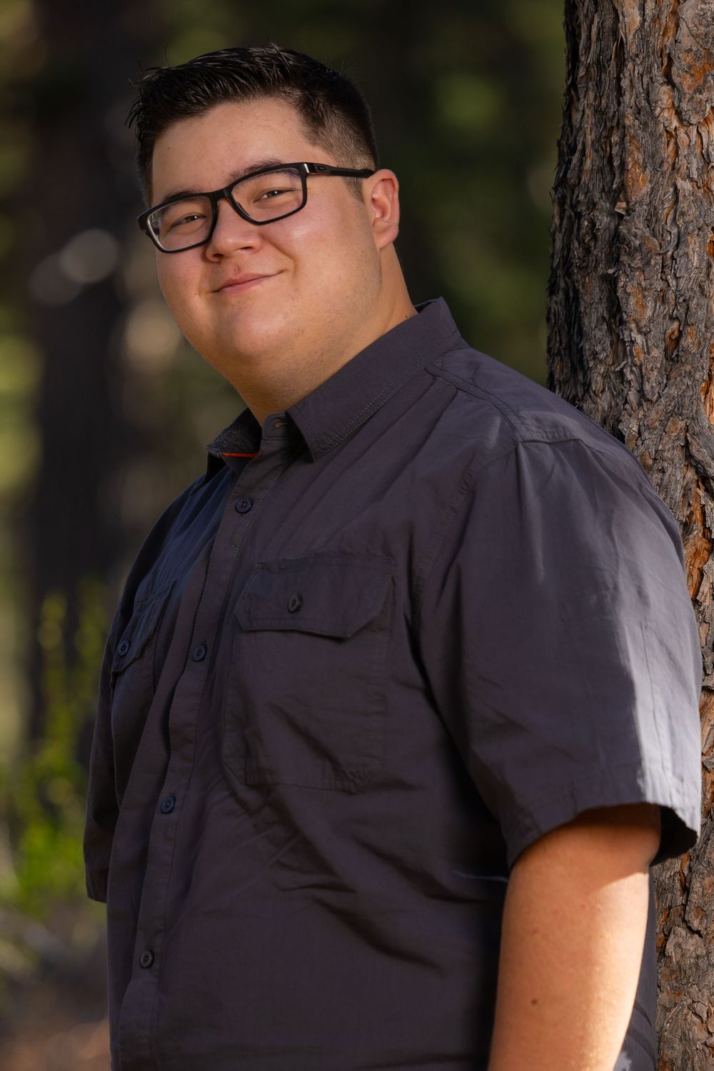 Person wearing glasses and a dark button-up shirt poses casually against a rustic tree trunk in an outdoor setting.