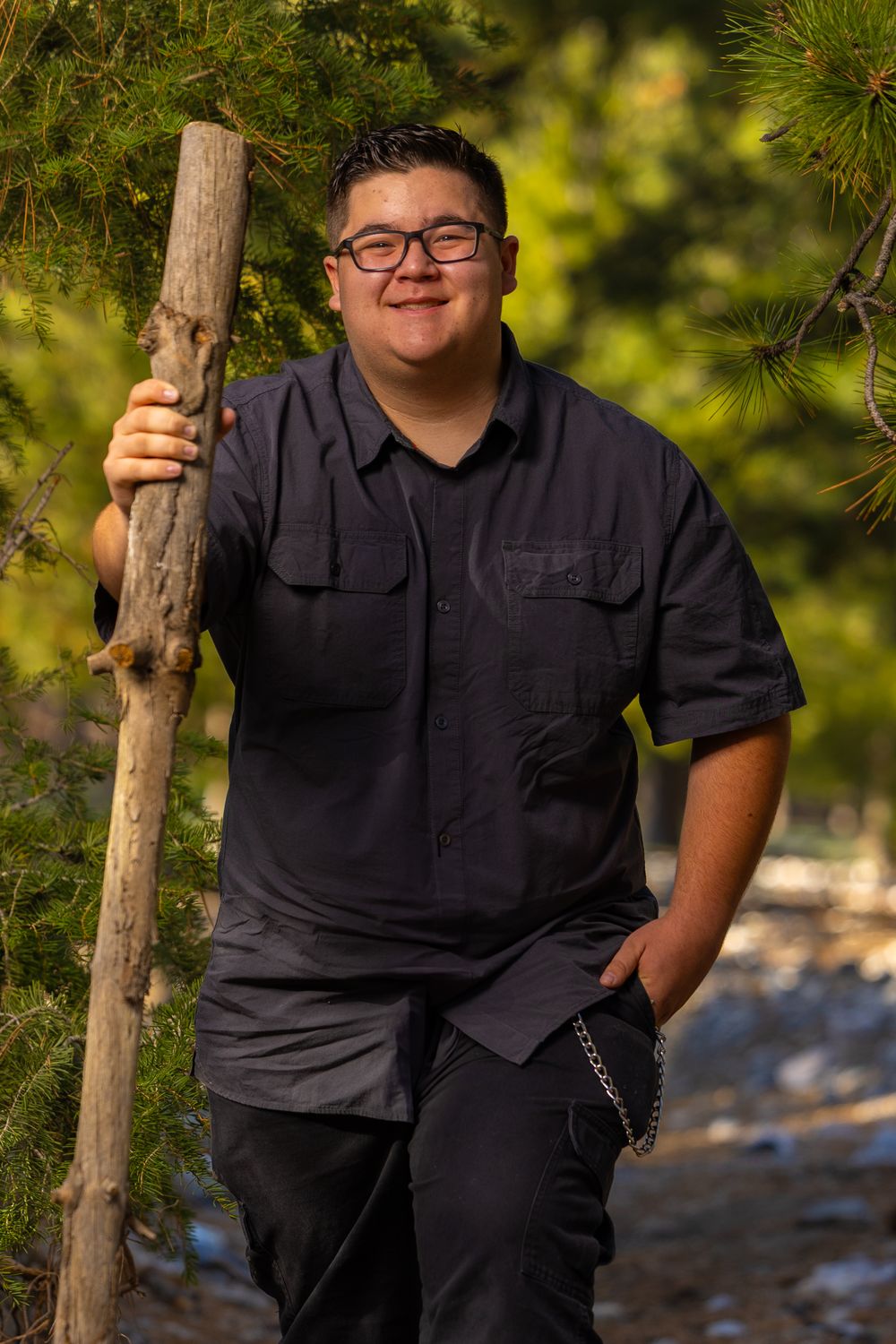 Someone in a black button-up shirt poses with a walking stick on a nature trail with pine trees in the background.