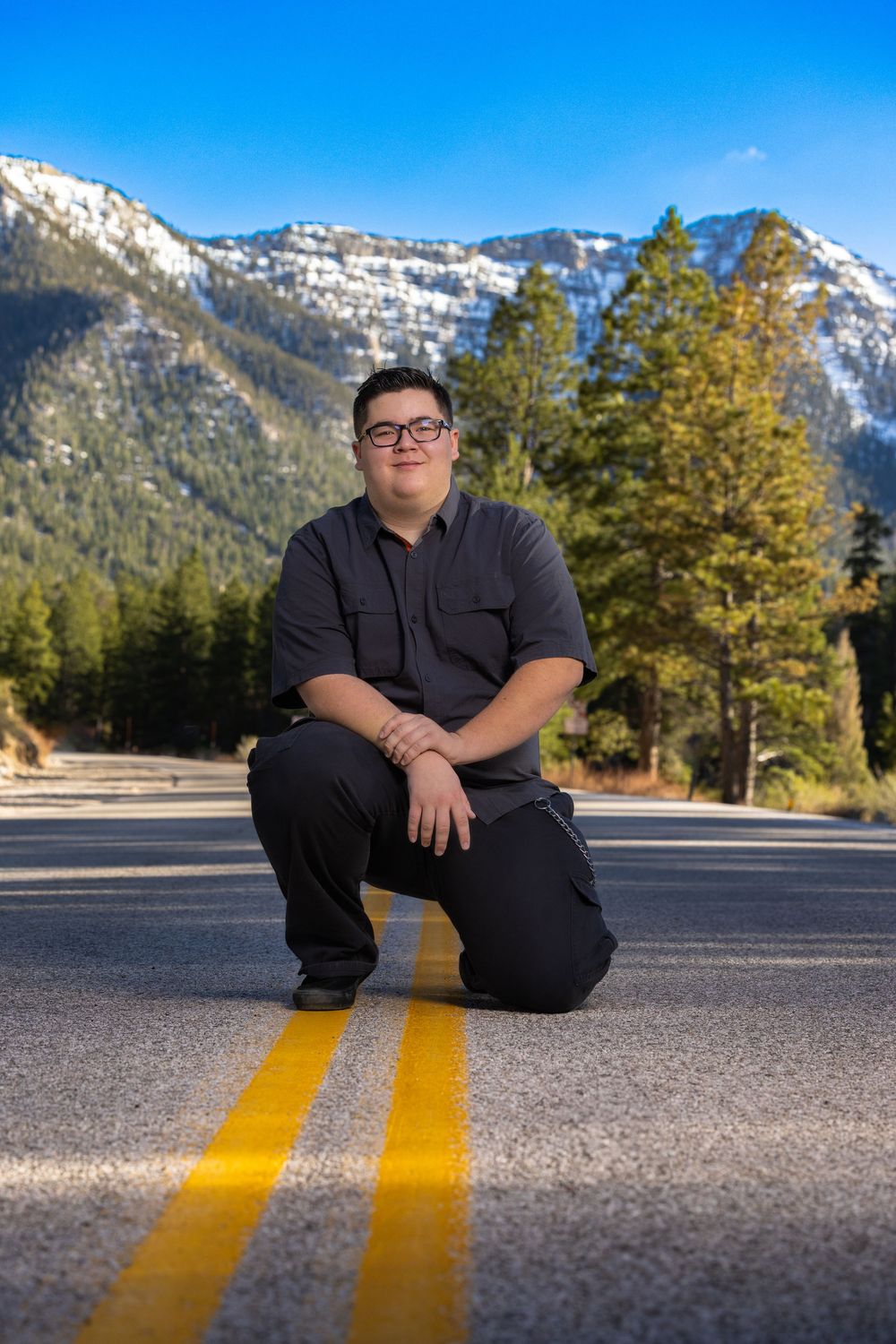 A lone figure kneels on a mountain road with snow-capped peaks and pine trees in the background on a sunny day.