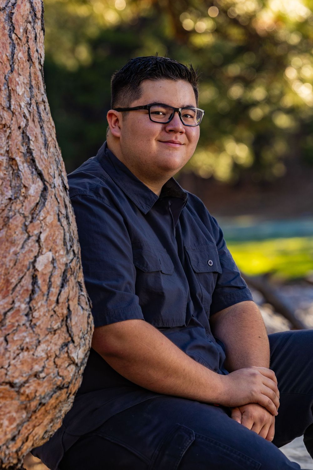 Portrait photo of someone in a blue button-down shirt sitting against a tree trunk in warm sunlit outdoor setting.