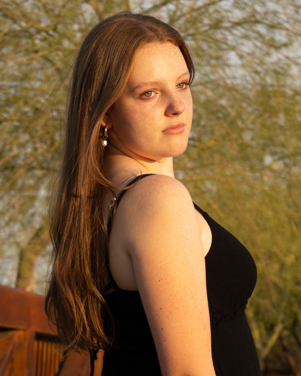 A figure in a black sleeveless dress poses against a blurred backdrop of trees during golden hour lighting.