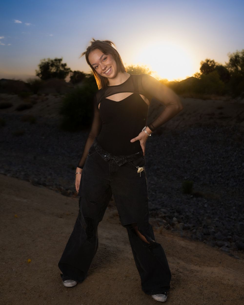 A person poses energetically against a sunset backdrop wearing all black attire on a desert path.