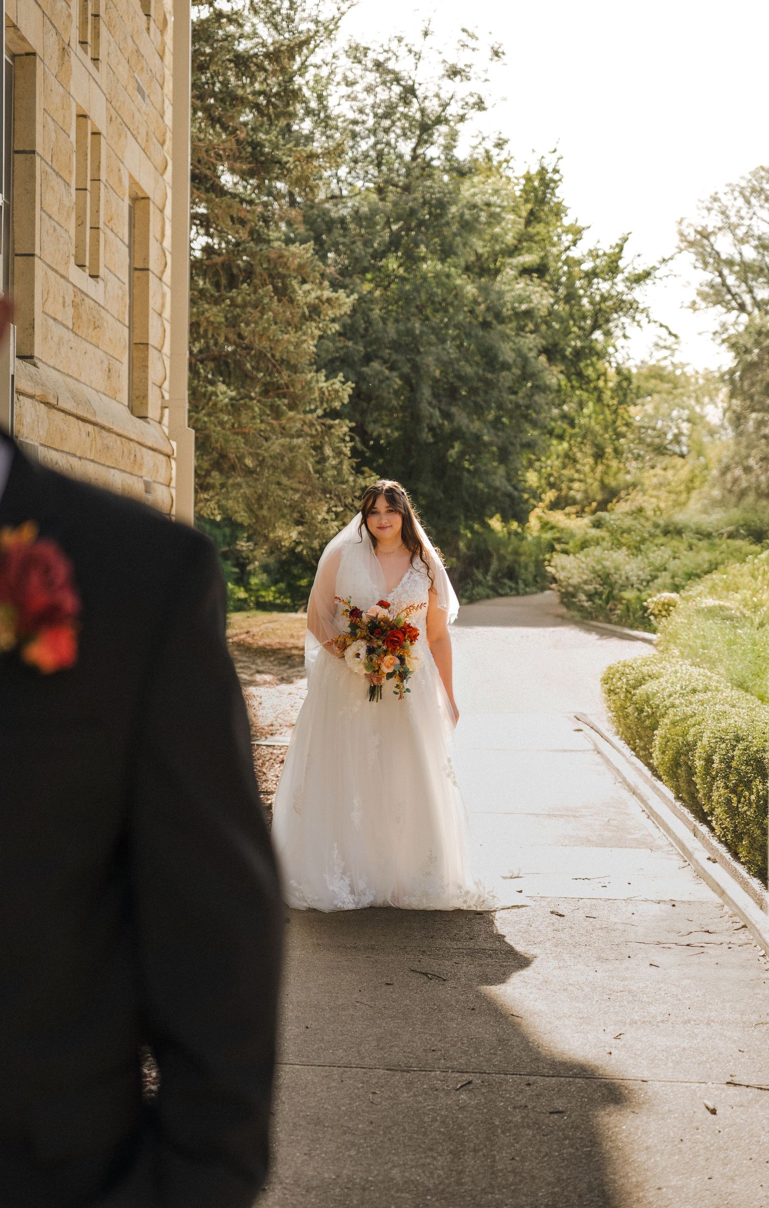 Someone in a flowing white wedding dress holding an autumn-colored bouquet stands on a stone path surrounded by greenery.
