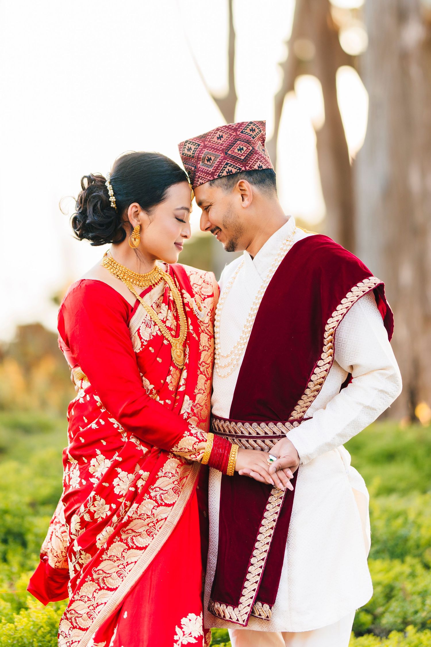 A couple in traditional red and burgundy wedding attire share a romantic moment outdoors at sunset.