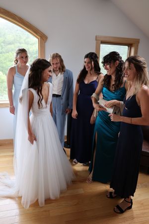 Wedding party members share an intimate moment getting ready in a sunlit room with wood trim and windows.