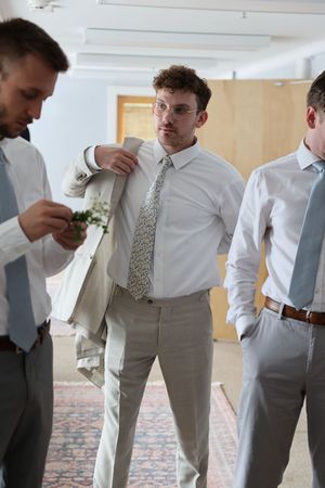 Groomsmen in light gray suits and white shirts adjusting their ties while getting ready for a wedding.