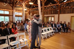 Two people embrace during a wedding ceremony in a rustic barn venue with string lights and wooden beams.