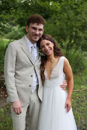 A happy couple poses outdoors on their wedding day wearing a light gray suit and flowing white wedding gown.