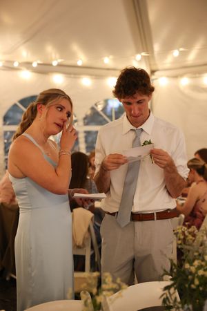 A couple shares an emotional moment while reading wedding speeches under a tent with string lights.