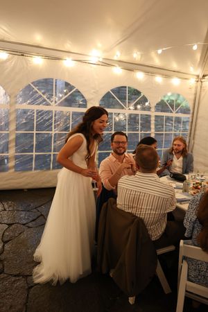 A reception under a white tent with twinkling string lights where guests gather at tables during what appears to be a celebration.