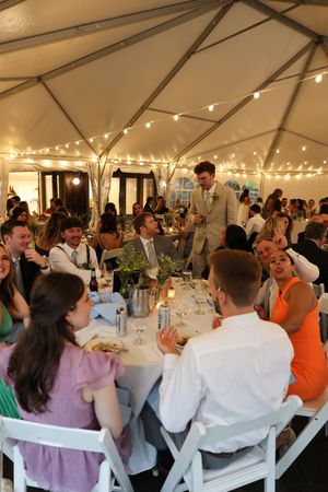 Wedding reception under tent with string lights as guests gather at tables for dinner service.