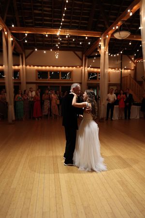 A couple shares a romantic first dance in a rustic barn venue decorated with string lights on the wooden beams.