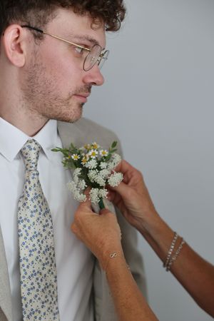 A wedding boutonniere made of small white and yellow flowers is being pinned to a light gray suit jacket.