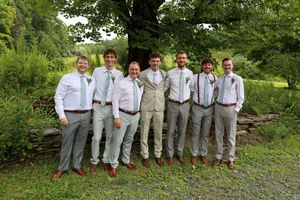 A group in matching gray pants and white shirts with suspenders pose together on a gravel path surrounded by greenery.