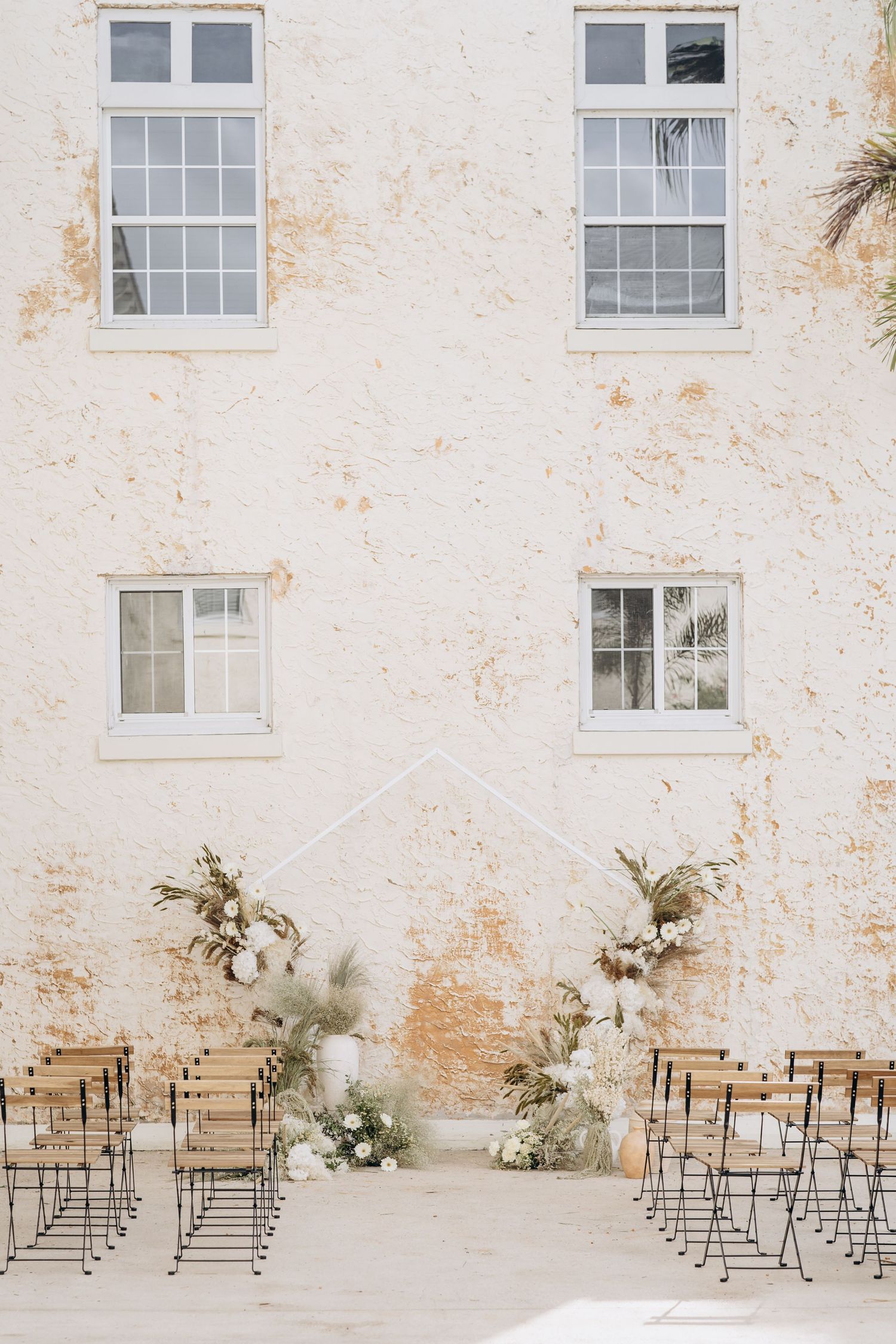 Minimalist wedding ceremony setup with metal chairs against a weathered white stone wall.