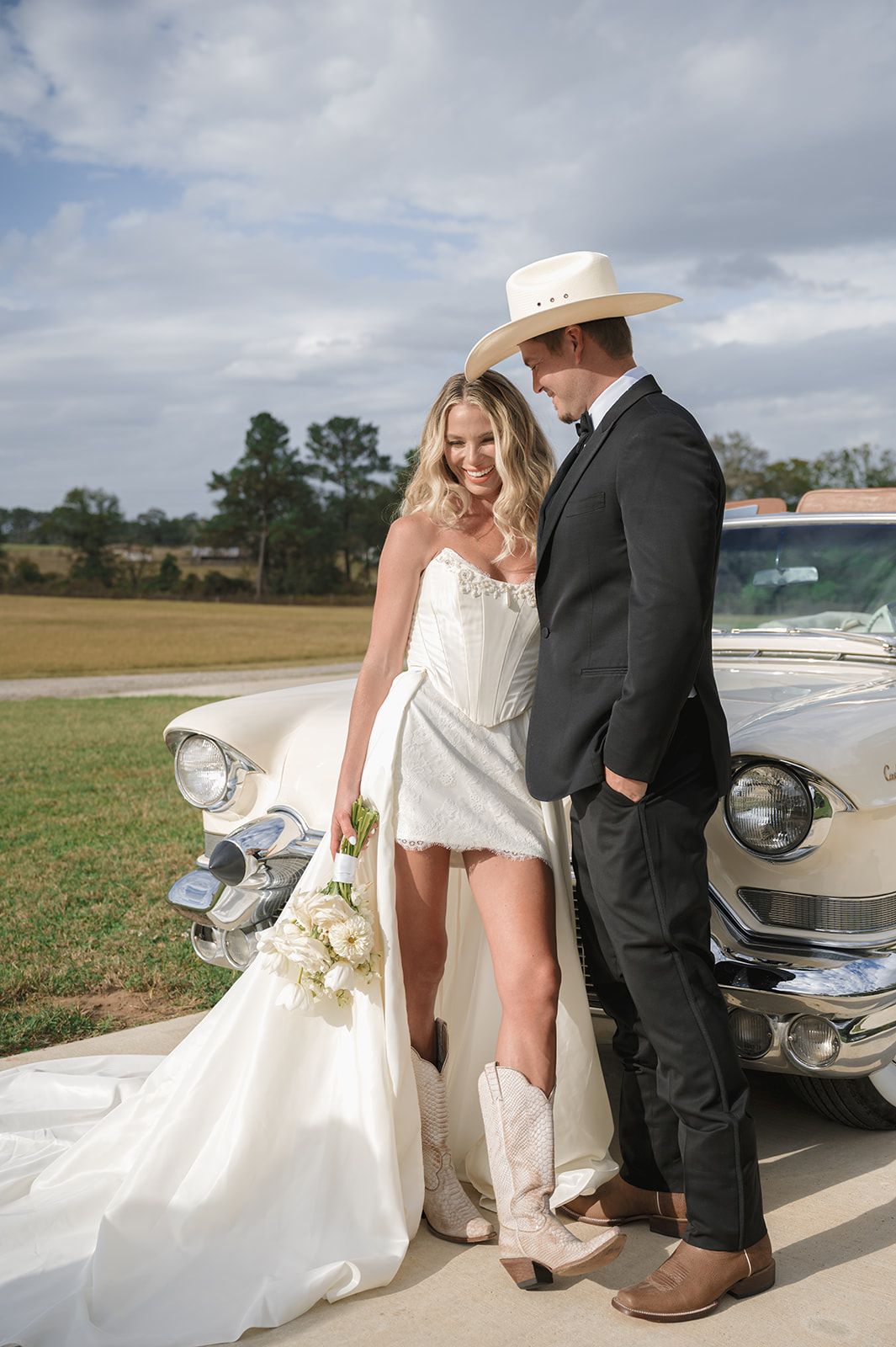 A bride and groom pose together near a vintage car at a rustic country wedding showing off her cowboy boots.