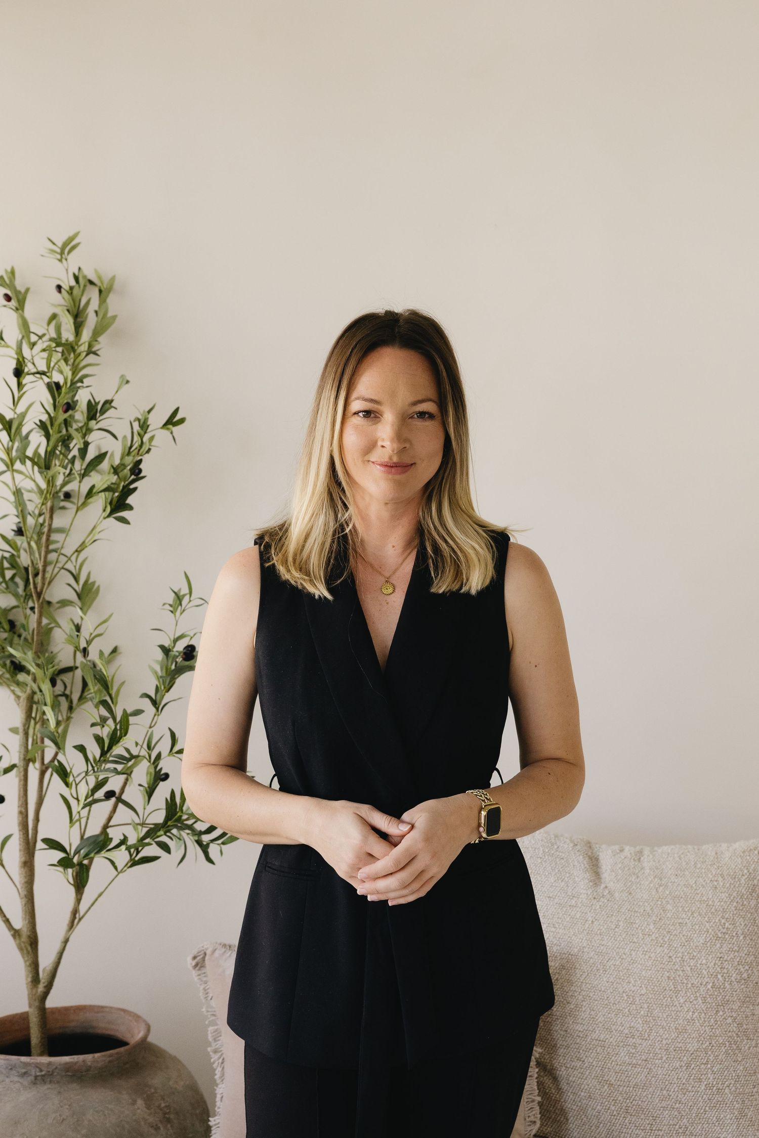Professional lifestyle portraits of a person in a black sleeveless dress seated on a beige couch with green plants in the background.
