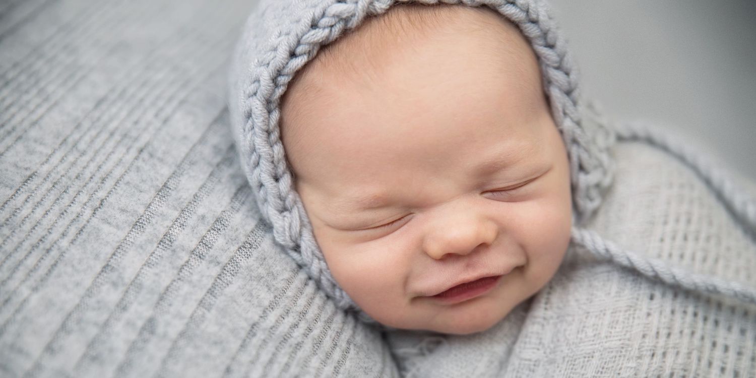 Newborn baby wearing grey knit hat sleeping peacefully on soft grey blanket.