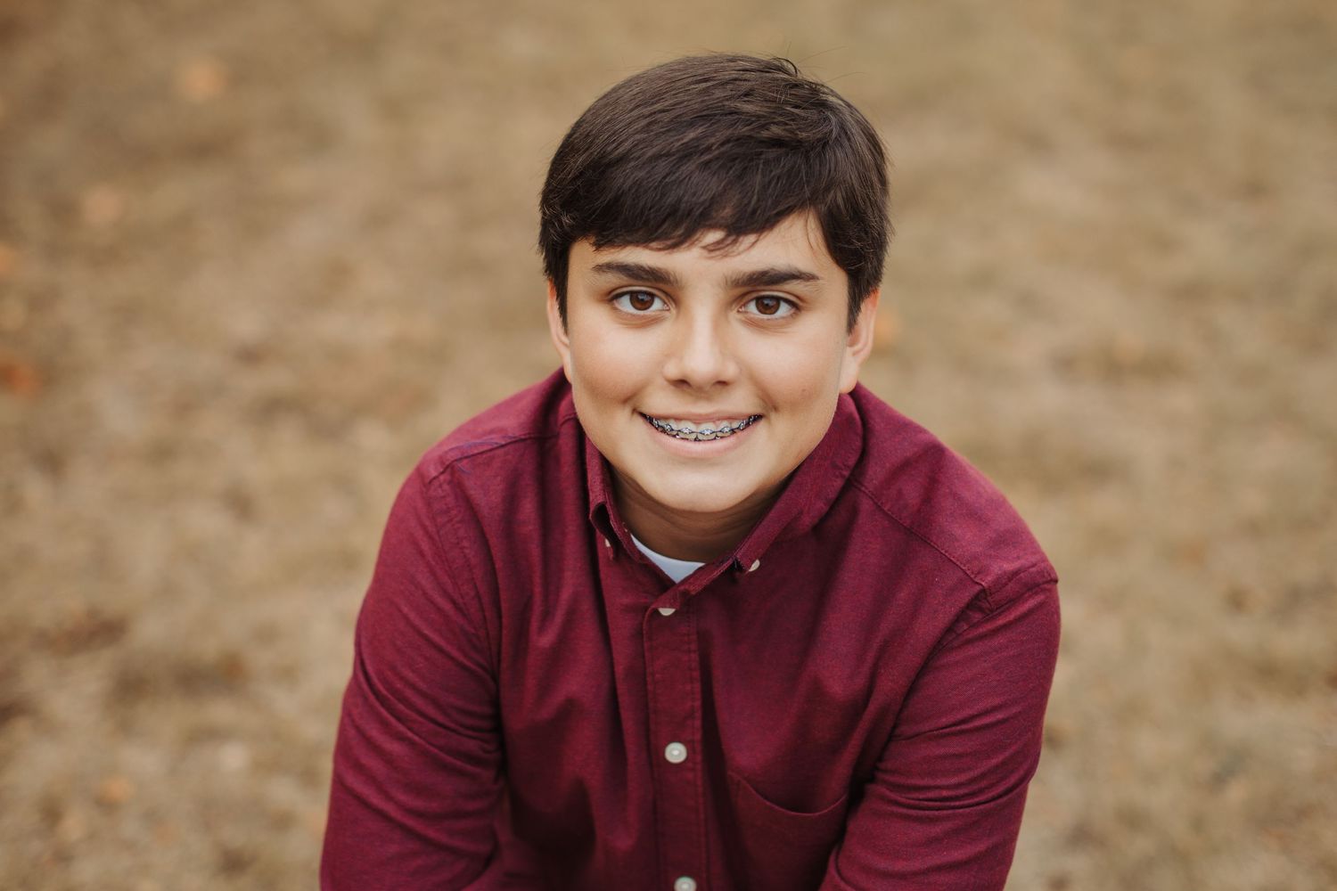 Someone wearing a maroon button-up shirt smiles warmly at the camera against a blurred outdoor background.