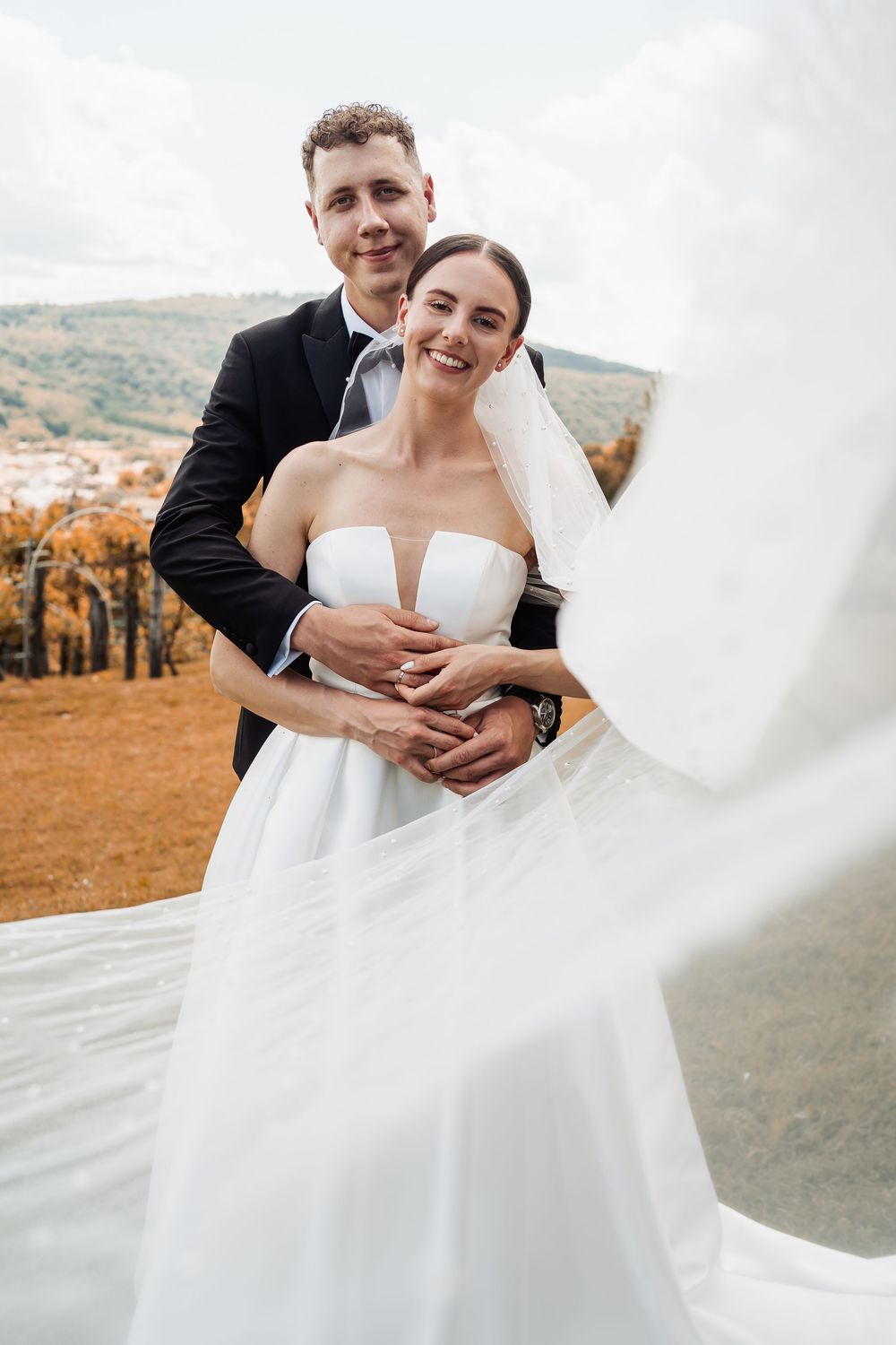A romantic outdoor wedding photo shows a couple embracing on an autumn hilltop as the bride's white dress flows in the wind.
