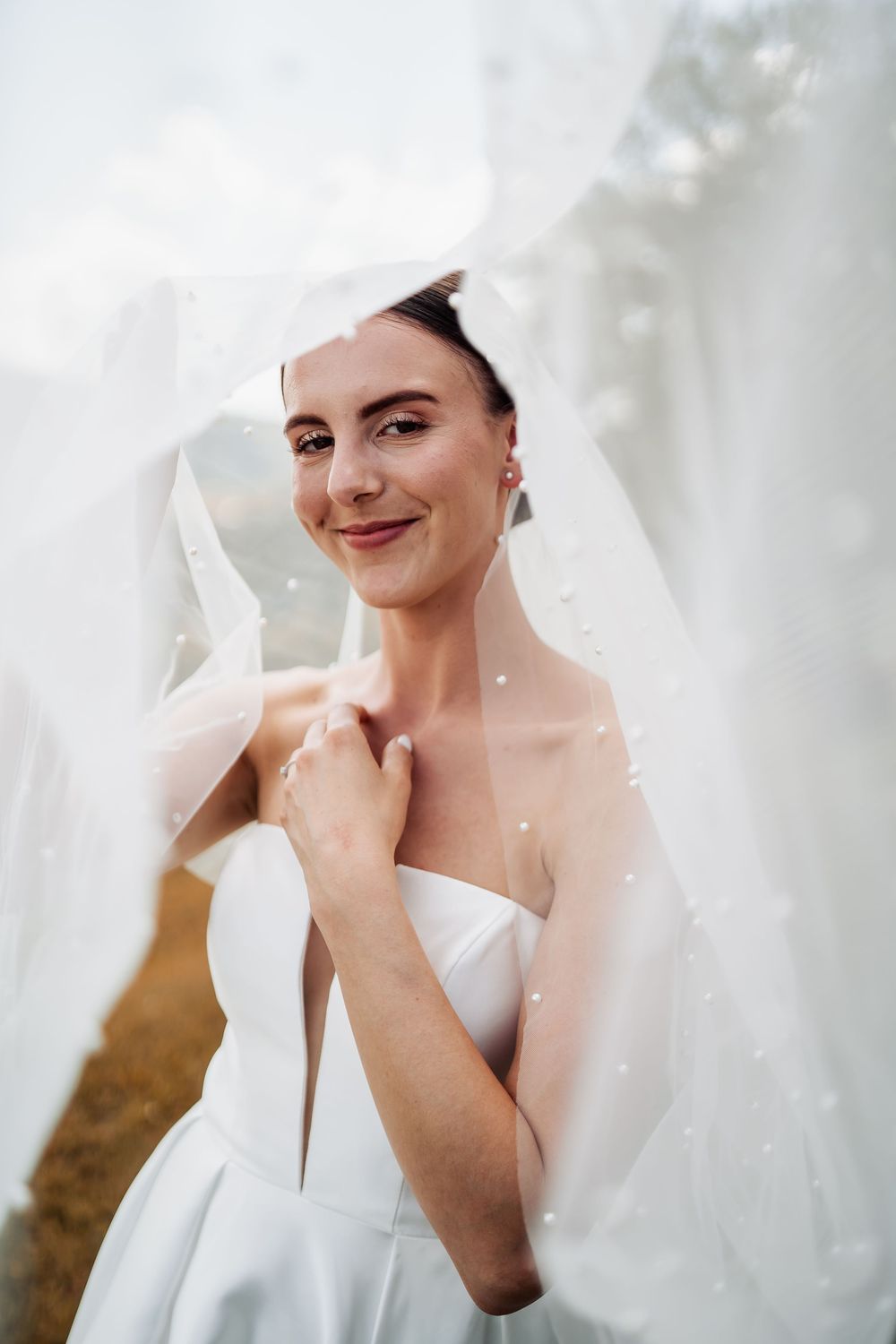 A bride in a classic white strapless wedding dress poses with a delicate veil on a soft, dreamy background.