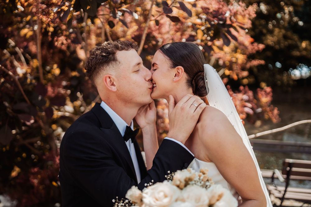 A romantic wedding couple shares a tender kiss outdoors surrounded by autumn-colored foliage and holding a white bouquet.