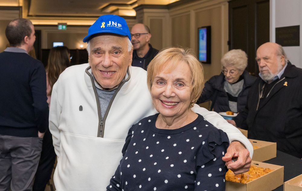 Two seniors smile together at a social gathering while wearing casual attire indoors.