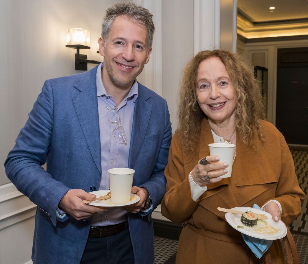 Two people share coffee and refreshments at an indoor event with a well-lit wall behind them.