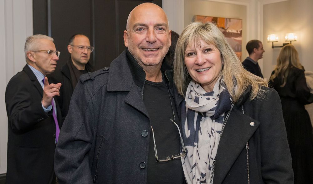 Attendees at an indoor gallery event wearing black coats and scarves in a warmly lit room.