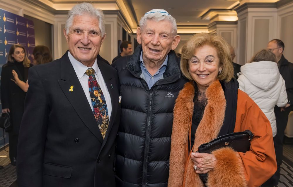 Three people stand together at an indoor event, with one wearing a business suit, another in a puffy vest, and one in orange.