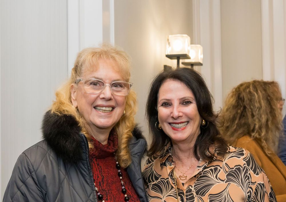 Two women smile warmly at a social gathering, with one wearing a fur-trimmed coat and the other in patterned attire.