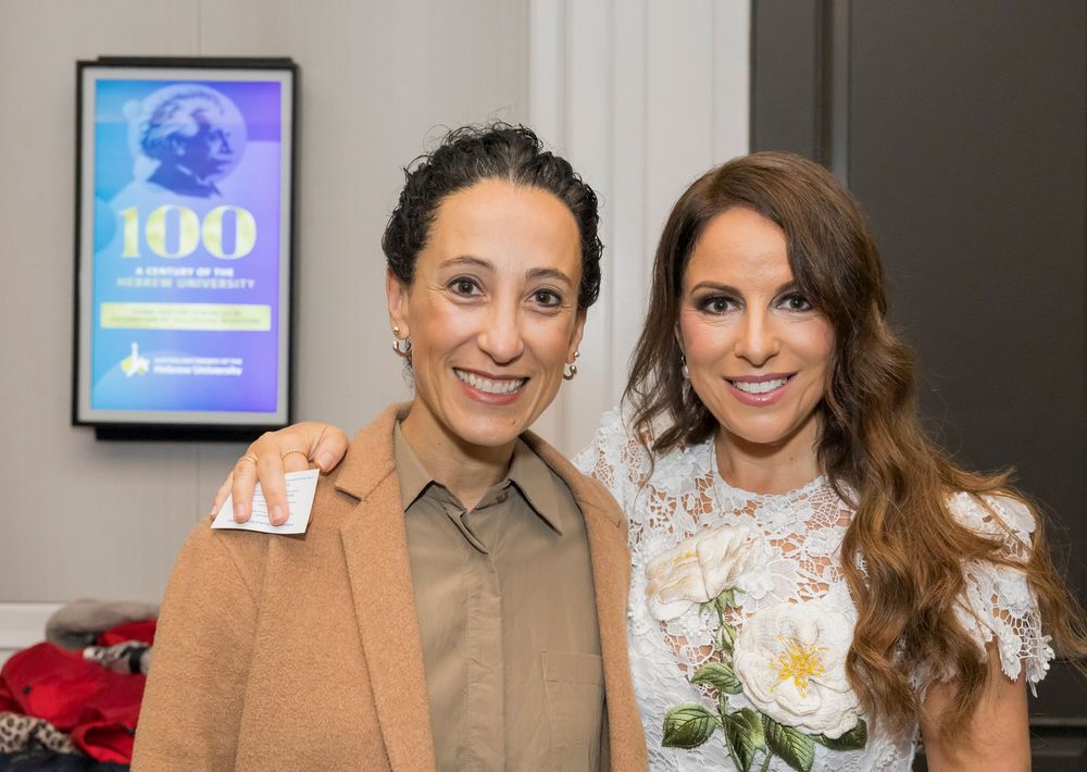 Two people pose together at an event with a digital display showing '100' visible in the background.