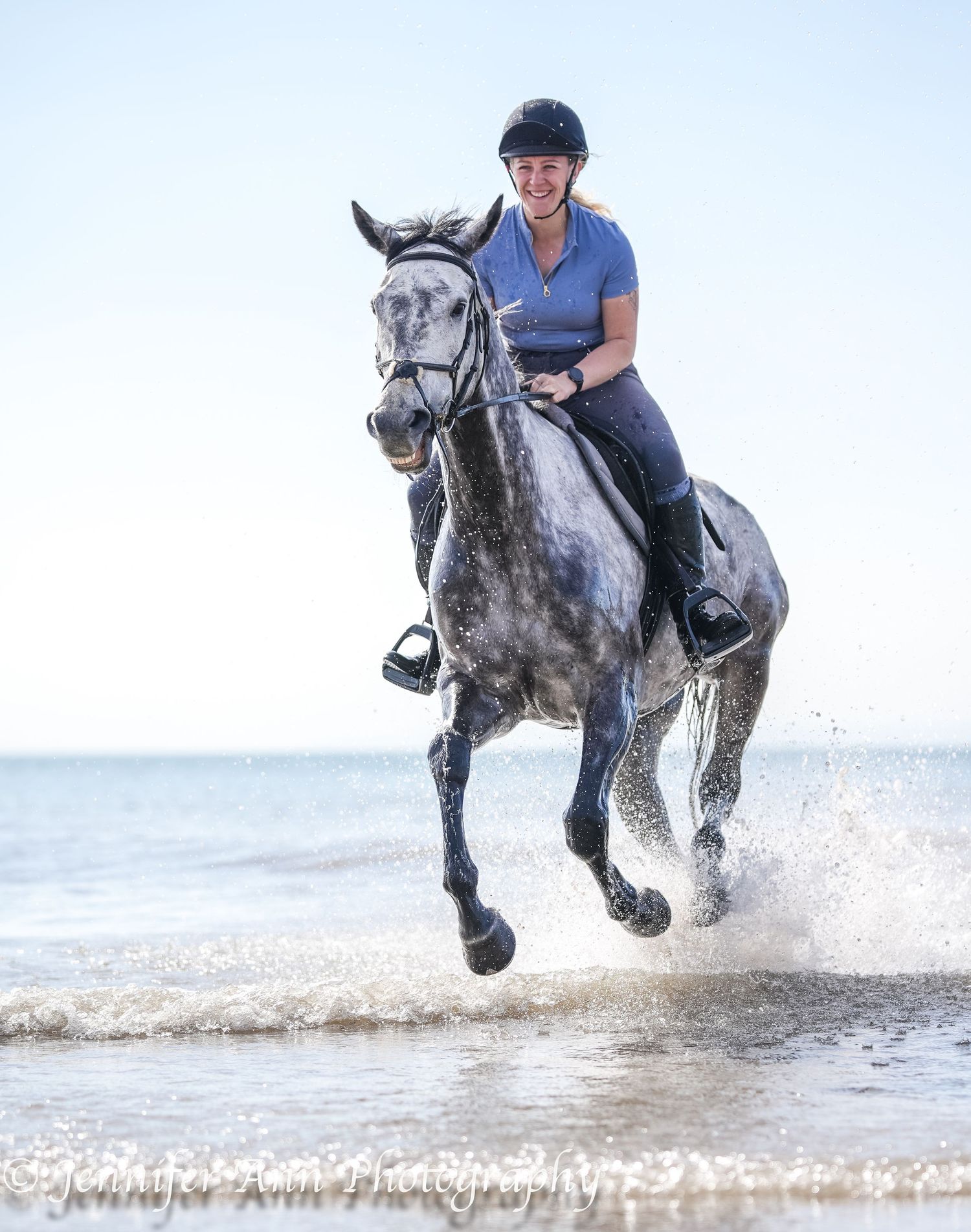 A dappled grey horse splashes through ocean waves near the shore.