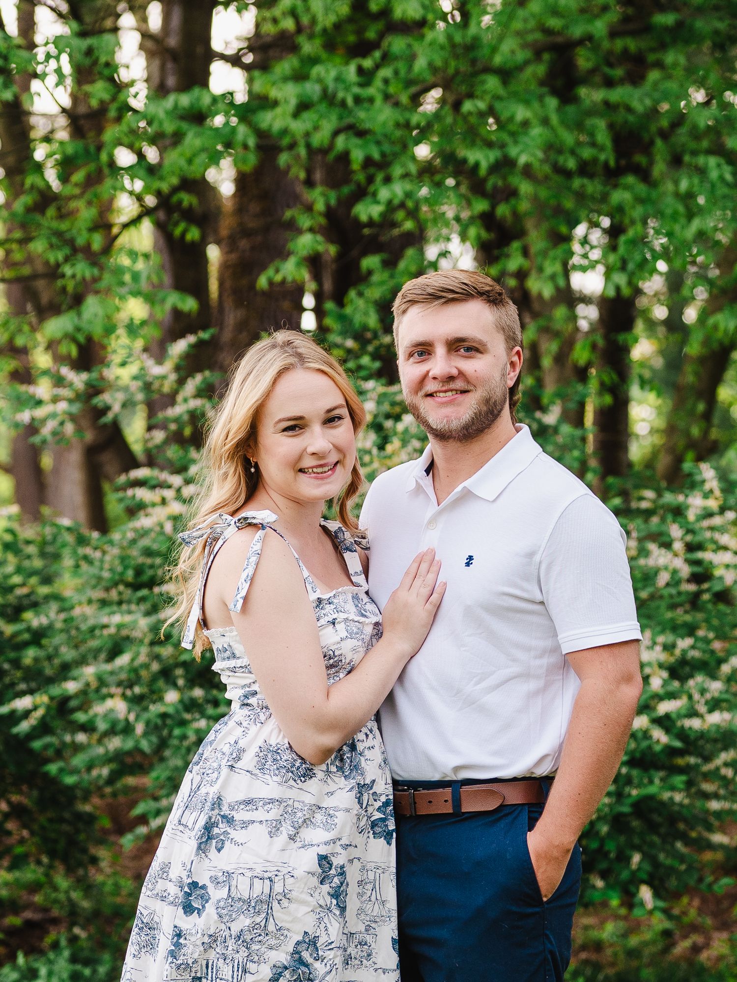 A couple poses together in a lush green forest in North Carolina wearing a floral dress and white polo shirt.